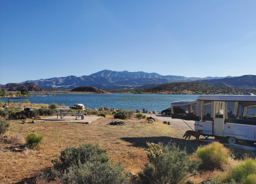 A serene lakeside campsite with mountains in the background, featuring a trailer and picnic area under a clear blue sky.